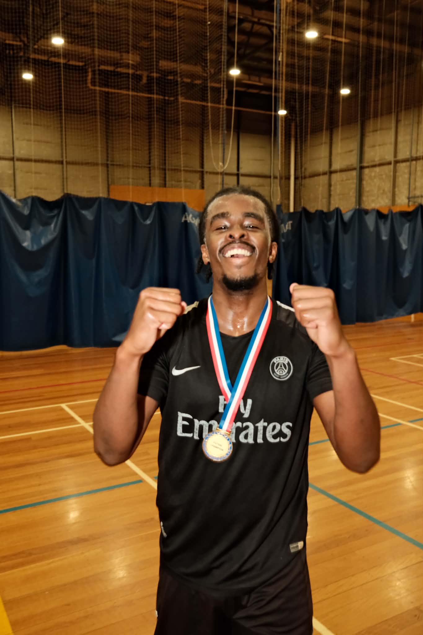 A smiling player wearing a medal raises both fists on the indoor court.